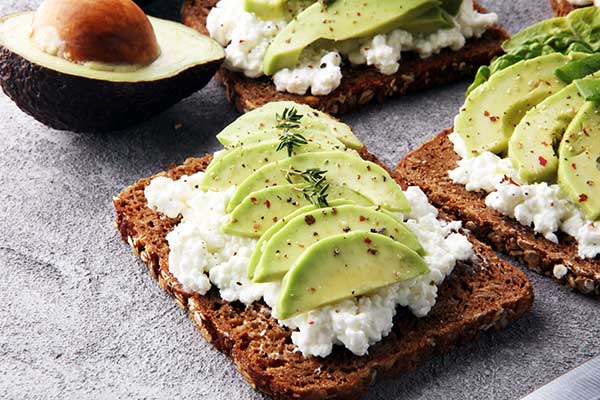 Tostada integral untada con queso crema y lonchas de aguacate por encima, salpimentadas con pimienta multicolor y una ramita de tomillo